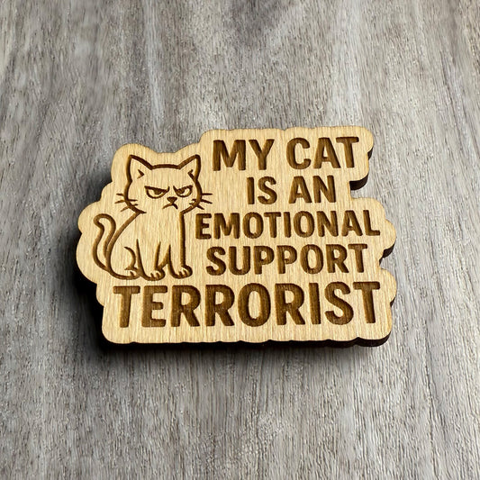 Angled view of the same engraved magnet reading “MY CAT IS AN EMOTIONAL SUPPORT TERRORIST” with an angry cartoon cat next to the text. The engraving has depth and clarity, with side lighting accentuating the wood grain and shadows.
