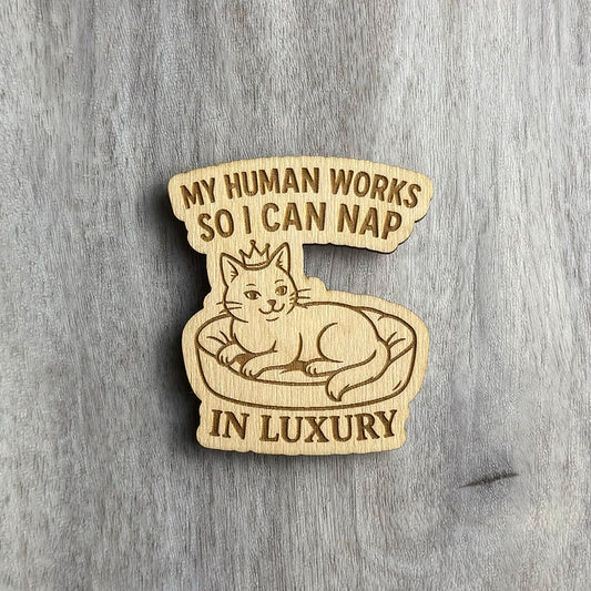 Laser-engraved wooden magnet with the phrase “MY HUMAN WORKS SO I CAN NAP IN LUXURY.” A smug cat wearing a crown lounges in a plush bed, engraved in a line-art style. The shape of the magnet curves around the cat and the stacked text layout.