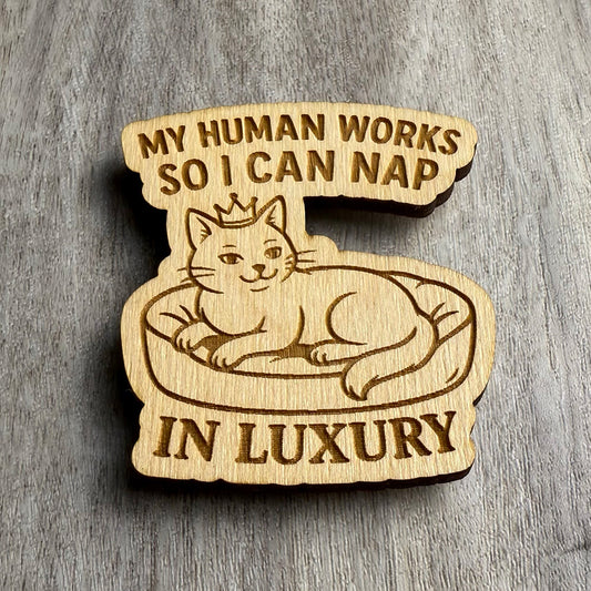Angled view of the engraved wooden magnet displaying “MY HUMAN WORKS SO I CAN NAP IN LUXURY,” with a crowned cat relaxing on a cozy pet bed. Lighting from the side highlights the engraved detail and cutout depth on the wood surface.