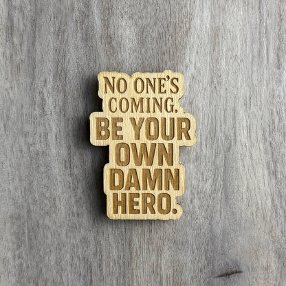 Laser-engraved wooden magnet with the empowering phrase “NO ONE’S COMING. BE YOUR OWN DAMN HERO.” The text is arranged in a tall stacked format using a mix of uppercase serif and bold block fonts. The shape of the magnet follows the rectangular structure of the layout.