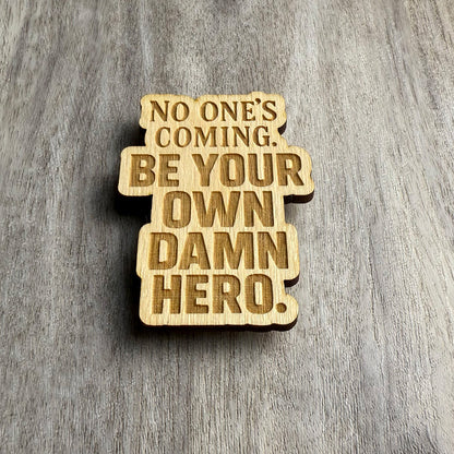 Angled view of the engraved wooden magnet that reads “NO ONE’S COMING. BE YOUR OWN DAMN HERO.” The lighting brings out the engraved depth and natural wood grain, adding emphasis to the bold typography.