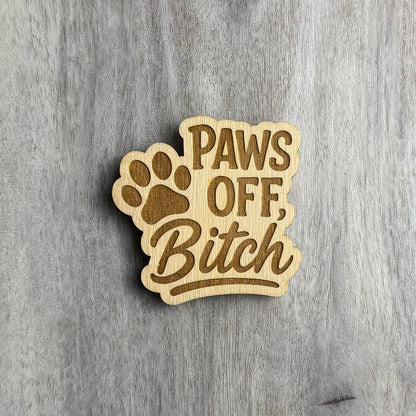 Laser-engraved wooden magnet with the phrase “PAWS OFF, Bitch” in a combination of bold uppercase and flowing script fonts. A large engraved paw print sits behind the word "PAWS," integrated into the design. The magnet has a playful cutout shape that follows the text and paw outline.
