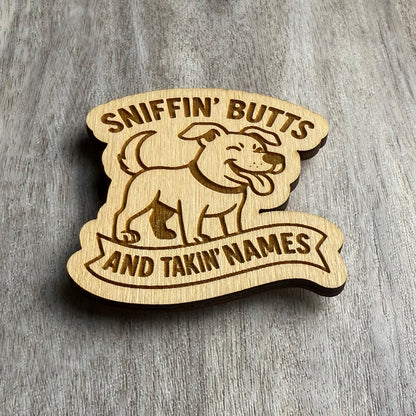 Angled view of a wooden magnet featuring a joyful cartoon dog above the phrase "SNIFFIN' BUTTS AND TAKIN' NAMES." The text is split across the top and bottom, engraved in bold font within a stylized banner. The magnet sits on a textured wood surface with natural grain.