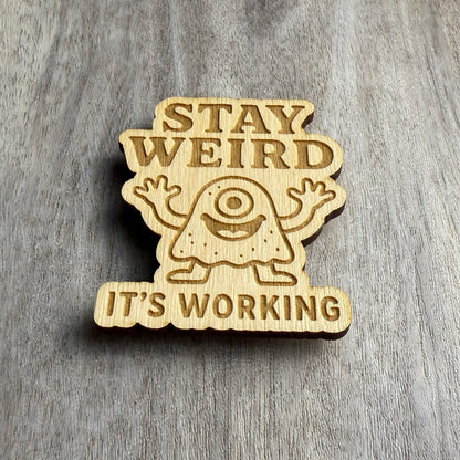 Angled view of a wood magnet featuring the phrase "STAY WEIRD IT'S WORKING" and a central image of a smiling cyclops monster with arms up and happy energy. The engraved design is cut into a wavy-edged shape, resting on a grayish wood background.