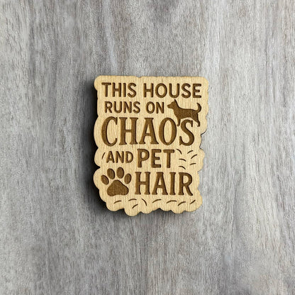 Wooden magnet engraved with the phrase "THIS HOUSE RUNS ON CHAOS AND PET HAIR." The design includes a silhouette of a dog at the top, a large paw print near the bottom, and scattered engraved pet hairs around the text. The border is scalloped and irregular, matching the playful messiness of the message. Background is a light gray wood surface.