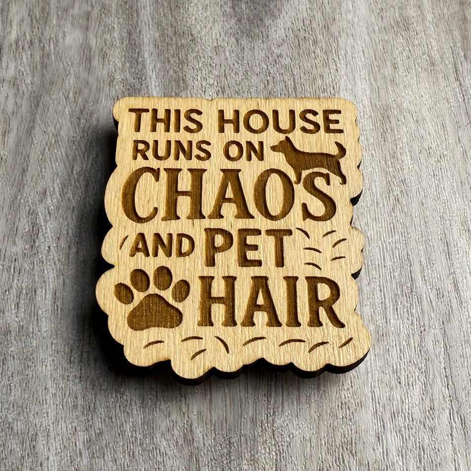 Angled view of a wood magnet engraved with the phrase "THIS HOUSE RUNS ON CHAOS AND PET HAIR," surrounded by scattered hairlines, a dog silhouette, and a large paw print. The magnet features a wavy, organic cutout shape and is placed on a textured wood grain surface.