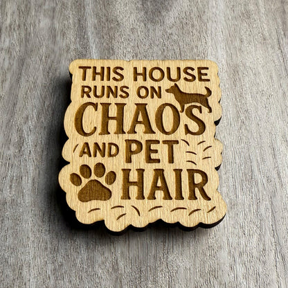 Angled view of a wood magnet engraved with the phrase "THIS HOUSE RUNS ON CHAOS AND PET HAIR," surrounded by scattered hairlines, a dog silhouette, and a large paw print. The magnet features a wavy, organic cutout shape and is placed on a textured wood grain surface.