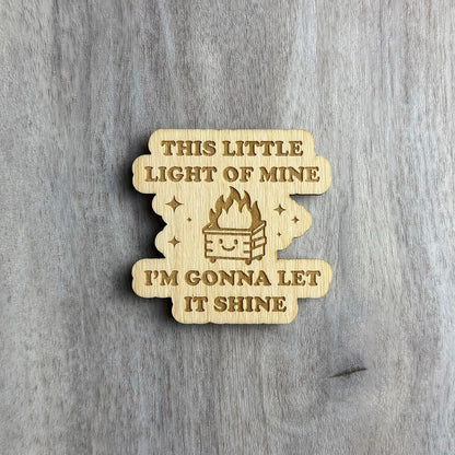 Wooden magnet engraved with the phrase "THIS LITTLE LIGHT OF MINE I'M GONNA LET IT SHINE" in uppercase letters. In the center is a smiling dumpster with flames coming out of it, surrounded by engraved sparkles. The shape has soft, cloud-like curves and is placed on a light gray wood surface.