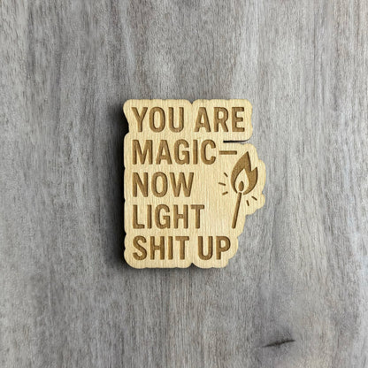 Wooden magnet engraved with the bold, all-caps phrase "YOU ARE MAGIC — NOW LIGHT SHIT UP." To the right of the text is an engraved illustration of a lit match with stylized flame lines. The magnet has a jagged, organic outline and is displayed on a gray-toned wood background.