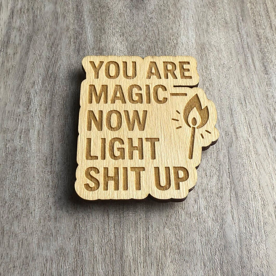 Angled view of a wooden magnet featuring the phrase "YOU ARE MAGIC — NOW LIGHT SHIT UP" in uppercase letters, with a burning match icon on the right. The engraved design is cut into a cloud-like wood shape and placed on a textured wood surface.