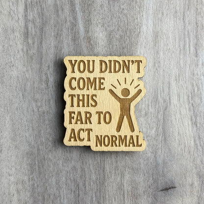Wooden magnet engraved with the phrase "YOU DIDN’T COME THIS FAR TO ACT NORMAL" in all caps. The right side features a figure with arms raised and radiating lines above its head, symbolizing energy or celebration. The magnet has an irregular, wavy border and is placed on a light gray wood surface.