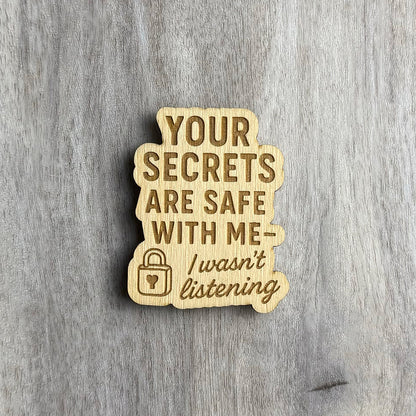 Wooden magnet engraved with the phrase "YOUR SECRETS ARE SAFE WITH ME — I wasn't listening" in a mix of bold block and cursive fonts. A small lock icon with a heart-shaped keyhole is engraved on the lower left. The magnet has a soft, bubbly border and is placed on a light gray wood background.