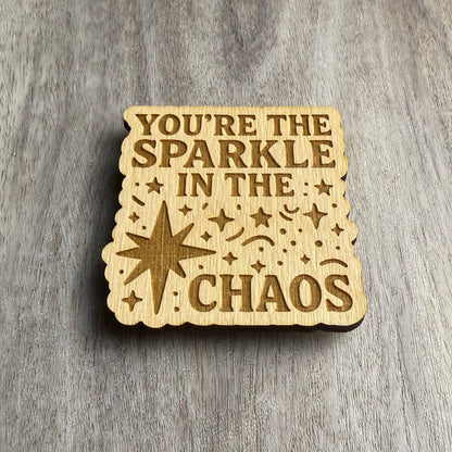 Angled view of a wood magnet featuring the engraved phrase "YOU’RE THE SPARKLE IN THE CHAOS" surrounded by scattered stars, confetti shapes, and a large starburst. The cutout has wavy, rounded edges and rests on a wood-textured background.