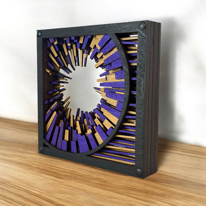 Metrorhythm V1 placed on a wood surface, catching light across its dimensional birch layers in black, purple, and gold.

